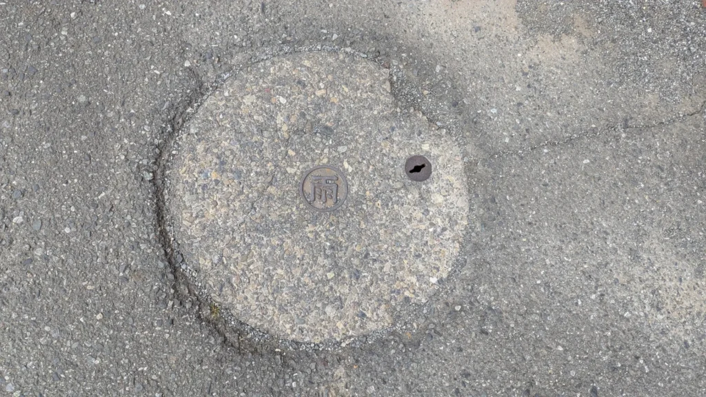 A manhole cover filled with asphalt to match the road, showing only a small rain emblem