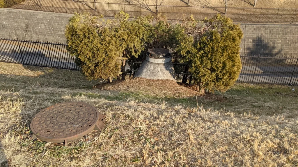 A manhole raised on a concrete cone on a grassy slope, with another cover lying on the ground in front