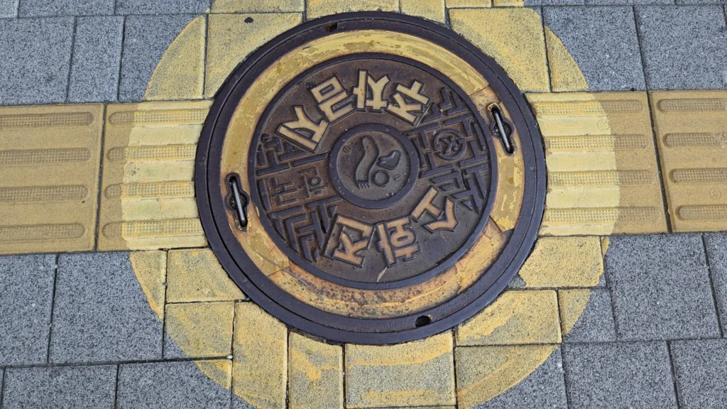 A bronze manhole cover with bold geometric lettering, set in yellow tactile paving