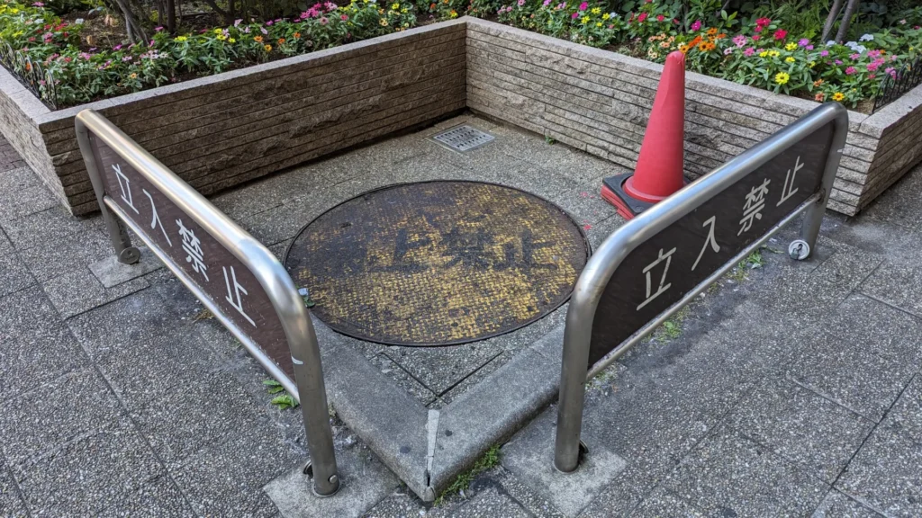 A manhole cover fenced off with No Entry barriers and a red traffic cone