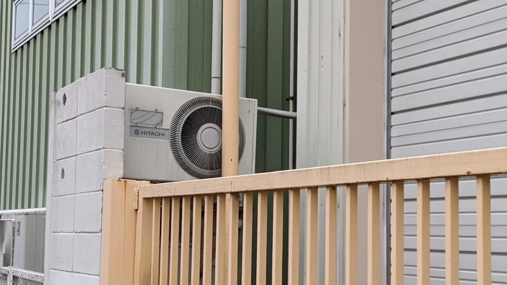 An old HITACHI right-eye outdoor unit on top of a block wall, peeking over a yellow fence