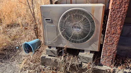 A weathered right-eye outdoor unit surrounded by dry grass next to a rusty pillar