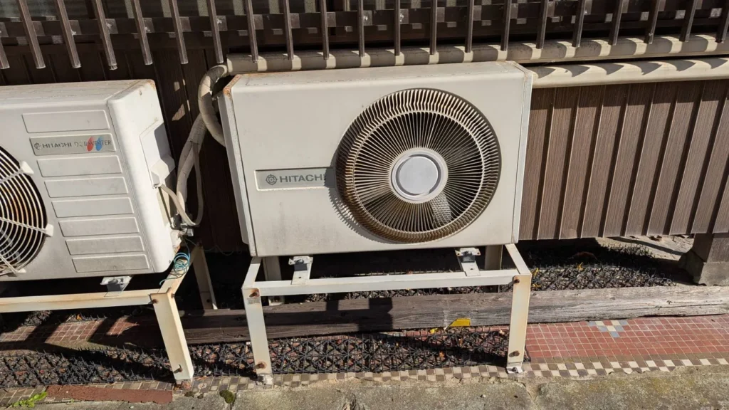 An old HITACHI right-eye outdoor unit on a metal stand next to a newer unit
