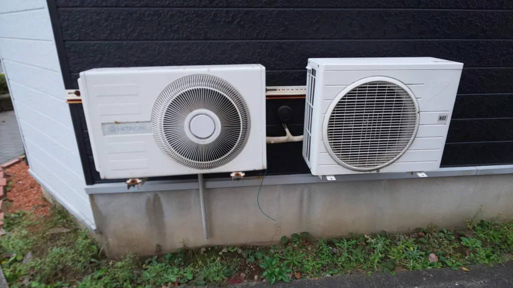 An old HITACHI right-eye outdoor unit beside a modern DAIKIN unit on a dark wall