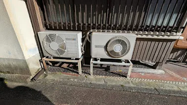 Two old outdoor units on metal stands along a narrow street